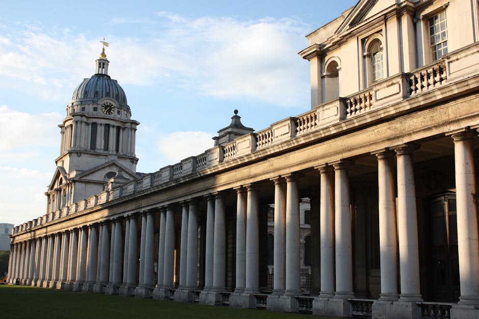 A historic building with a large central dome topped by a golden finial, featuring a clock on the front façade. The building has classical architectural elements such as columns, arches, and decorative stonework. An extensive colonnade with evenly spaced columns runs along the front of the structure, providing an open walkway beneath. The scene is illuminated by natural daylight, with blue skies and some clouds in the background. The building's surroundings include green grass and part of a paved area, indicating an outdoor setting. The photograph is taken from a slightly lower angle, emphasizing the building's grandeur. This image relates to house removals and home relocation services, capturing a prominent London landmark, and naturally supports content about moving logistics and furniture transport, especially within historic or significant architectural contexts provided by Man and Van Greenwich.