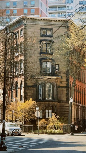 A multi-storey stone building situated at an urban street intersection, featuring three prominent arched and rectangular windows on each floor, with a decorative stone façade showing signs of weathering. The building is surrounded by leafless trees and overgrown bushes, indicating a mature, established property, with some yellow foliage visible at street level. To the left, part of another building with a brick exterior is visible, while to the right, an orange-hued building with rounded window frames can be seen. In the foreground, there is a pedestrian crossing with painted stripes, tram or trolleybus wires overhead, and a white van parked at the curb, suggesting active street use. The overall scene reflects a residential area within a city, where home relocation and furniture transport could involve navigating narrow streets and careful planning, as managed by local removal specialists such as Man and Van Greenwich.