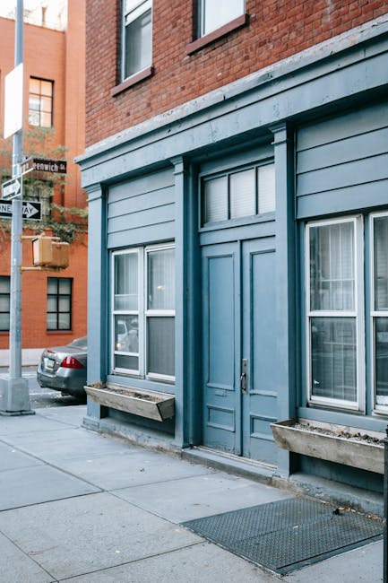 The image shows the exterior of a small building with a blue facade, featuring a central double door and three large windows, each with white frames and partially covered by closed blinds. Two stone window boxes without plants are mounted below the windows, and a wooden bench is positioned beneath the left window. The building's interior is not visible, but the scene suggests it is part of a residential or commercial property situated on a quiet urban street. A parked car is visible on the street to the left, and the sidewalk is clean with a metal grate near the entrance. The surrounding environment includes other brick buildings, indicating a city location, and the lighting appears to be natural daylight. This setting could relate to home relocation or furniture transport preparations, as part of a professional removals process conducted by Man and Van Greenwich, especially relevant for narrow street house removals in Greenwich.