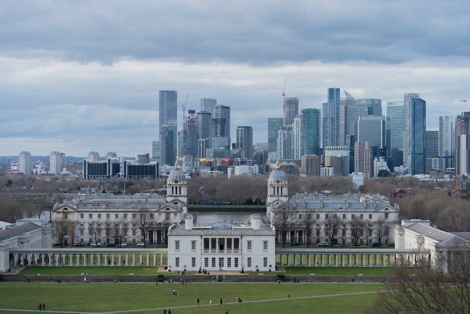 A wide view of a city skyline during daytime, featuring modern high-rise buildings with glass facades and a variety of architectural styles. In the foreground, there is a large green park area with pe