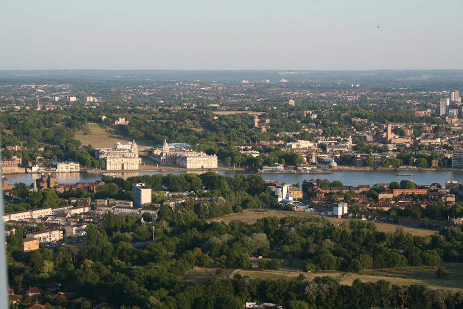 Aerial view of Greenwich, London, showing the River Thames flowing through the centre with historic and modern buildings on both sides, surrounded by lush green parks and residential areas. The scene is captured in daylight with clear weather, highlighting the extensive greenery, the detailed architecture of the Greenwich Palace ruins, and the broad cityscape stretching into the distance. This image illustrates the urban environment and scenic surroundings typical of London, relevant to house relocations and moving services such as those provided by Man and Van Greenwich, with the focus on efficient furniture transport and logistics in relocation projects within SE10 and Greenwich borough.