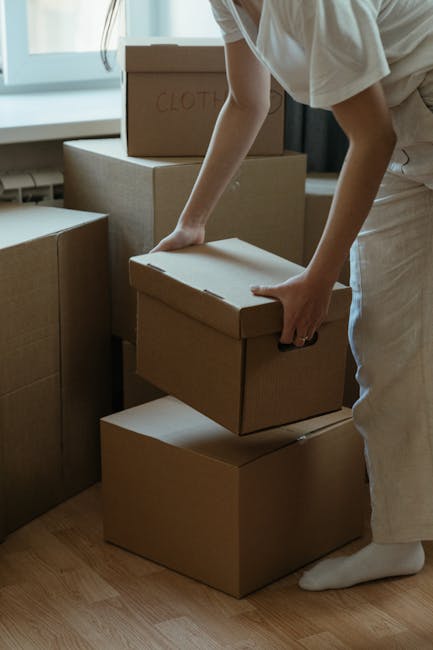 A person wearing light-colored clothing and white socks is seen inside a room, carefully placing a medium-sized cardboard box onto a stack of other packed boxes. The boxes are plain, brown, and mostly sealed with packing tape, some with handwritten labels such as 'clothes.' Natural daylight is coming through a nearby window, illuminating the scene. The environment appears organized for a home relocation or moving process, with several packed boxes arranged on a wooden floor, close to the window and near a doorway or interior wall. The person is engaged in packing or preparing items for moving, a typical part of furniture transport and home removals, as performed by company services like Man and Van Greenwich.