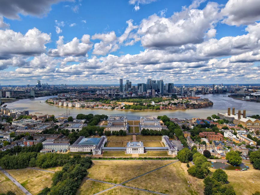 Aerial view of Greenwich, London, taken during daytime under partly cloudy skies, showing the River Thames curving through the city with residential and commercial buildings along its banks. In the foreground, there are green parks, open grassy areas, and pathways with some trees. The prominent Royal Observatory, with its neoclassical architecture and white facade, is situated on the hilltop surrounded by well-maintained gardens. Further in the background, the modern skyline of central London with skyscrapers and high-rise buildings stretch across the horizon. The scene captures a broad perspective of Greenwich’s historic and contemporary landscape, suitable for illustrating home relocation, furniture transport, or packing and moving activities near Cutty Sark and Greenwich Market.