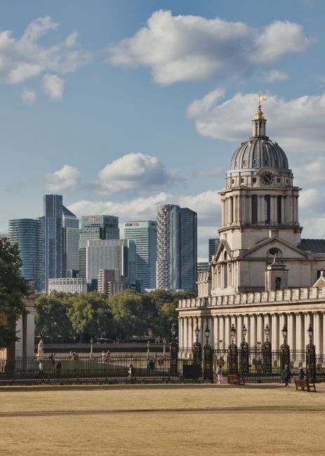 A view of a historic cathedral with a domed clock tower and ornate architectural details, situated in a park area with a black wrought-iron fence in the foreground. Behind the cathedral, there is a modern city skyline featuring tall glass skyscrapers, including recognizable office buildings with company logos. The park has a grassy lawn and several people walking or standing near the fence, with some sitting on benches. The sky is partly cloudy with patches of blue. This scene captures the contrast between traditional and contemporary architecture in Greenwich, relevant to house relocations and moving services by Man and Van Greenwich, with the city backdrop emphasizing urban moving logistics and the importance of professional packing and furniture transport.