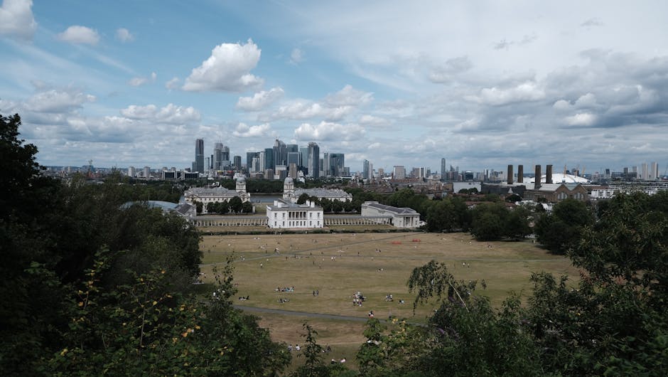 A wide view of Greenwich Park in London with a large open grassy area in the foreground, scattered with people relaxing and picnicking. Behind the park, the historic Queen's House with its white facade and classical architecture is visible, surrounded by trees. In the distance, the modern skyline of London's financial district with tall office buildings and skyscrapers rises under a partly cloudy sky. The scene is captured in daylight, emphasizing the contrast between the historic and contemporary elements of Greenwich. This setting illustrates typical surroundings for home relocation and furniture transport services provided by Man and Van Greenwich, showcasing the importance of careful planning, packing, and loading during moving processes within urban environments.