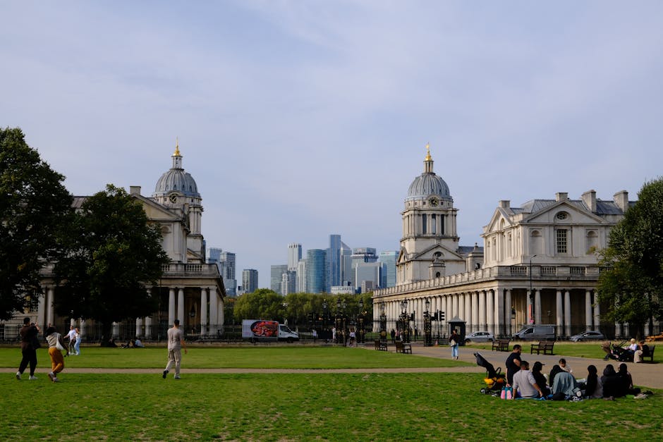 A wide view of a historic urban park with green grass and several people sitting on the lawn and walking along paved pathways. On the left and right sides of the image, there are large, classical-style buildings with white stone facades, columns, and domed rooftops topped with golden spires, indicative of the architectural style in Greenwich. In the background, a modern city skyline with tall glass skyscrapers is visible, contrasting with the historic buildings. The scene is set during daylight with clear weather, and a few trees flank the open space, providing partial shade. The park appears to be used for leisure and social gatherings, with some individuals pushing strollers, chatting, or relaxing on the grass. The overall atmosphere suggests a peaceful, urban setting suitable for home relocation or furniture transport activities, with the historic architecture emphasizing the importance of careful packing and moving processes typically coordinated by professional removals services like Man and Van Greenwich.