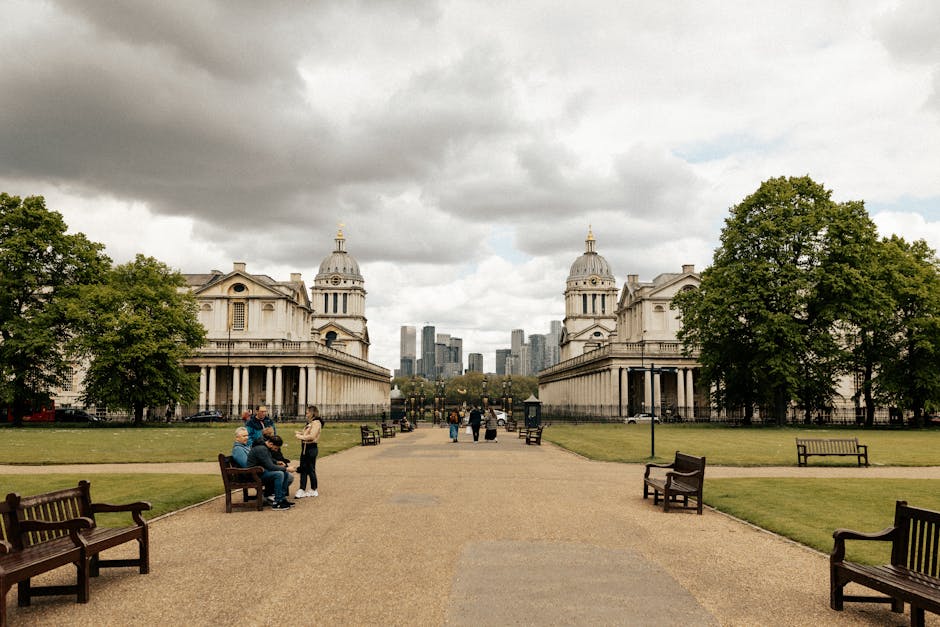 A wide view of Greenwich Park in London with a large open grassy area in the foreground, scattered with people relaxing and picnicking. Behind the park, the historic Queen's House with its white facade and classical architecture is visible, surrounded by trees. In the distance, the modern skyline of London's financial district with tall office buildings and skyscrapers rises under a partly cloudy sky. The scene is captured in daylight, emphasizing the contrast between the historic and contemporary elements of Greenwich. This setting illustrates typical surroundings for home relocation and furniture transport services provided by Man and Van Greenwich, showcasing the importance of careful planning, packing, and loading during moving processes within urban environments.